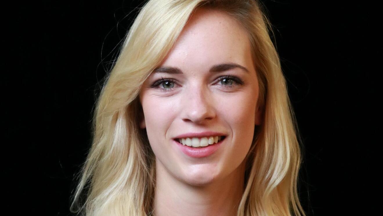 Eine Frau mit blonden langen Haaren und einem weißen T-shirt mit schwarzem Blaser schaut in die Kamera und lächelt.