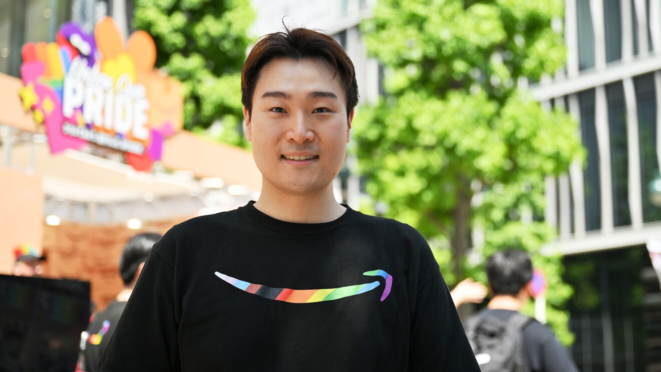 レインボーカラーのAmazonのロゴが入ったTシャツを着ている男性背景に茶色のブースとビル、街路樹が見える