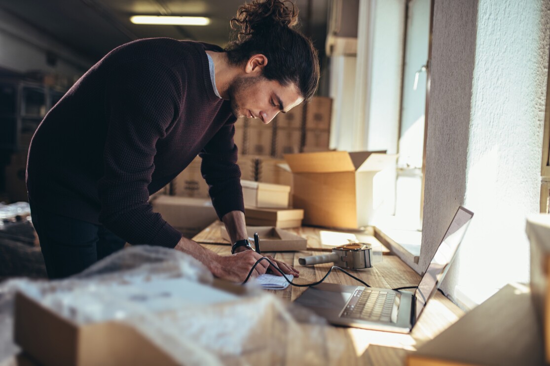 EIn Mann beugt sich über einen Schreibtisch, vor sich einen aufgeklappten Laptop, während im Hintergrund mehrere geöffnete Kartons zu sehen sind.