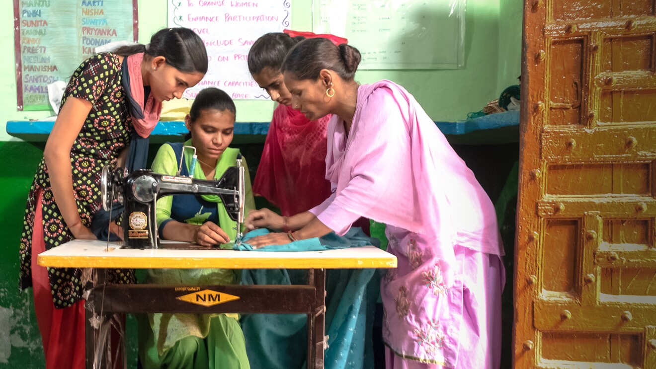 Three women stand around a woman who sits at a sewing machine.