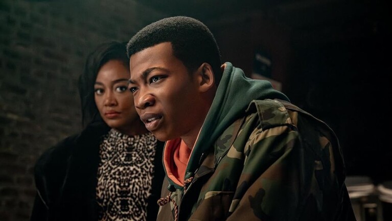Man in the foreground looking intensely to the side, and a woman standing in the background looking forward with a serious expression