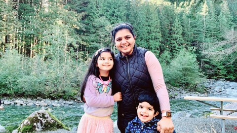 An image of a mother smiling for a photo with her two children in an outdoor area with trees and a creek behind them.