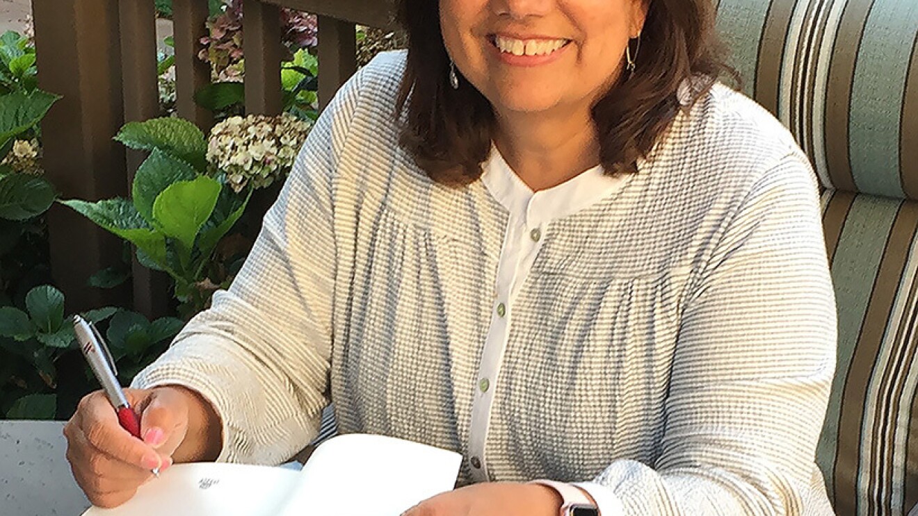Amazon author Barbara Freethy sits on a deck in the sunshine behind a stack of her books. She is autographing one book.