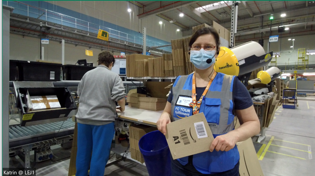 Katrin Stein, Tourleiterin im Amazon Logistikzentrum Leipzig, zeigt wie das Verpacken funktioniert. Sie trägt eine blaue Weste, ein Headset und einen Mundschutz, steht vor einer Verpackungsstation und hält einen braunen Amazon Umschlag in der Hand. Im Hintergrund sieht man Förderbänder und eine Mitarbeiterin am Verpackungstisch arbeiten.