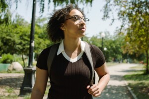 nicole, an amazon future engineer scholarship recipient, walks down a campus road while wearing a backpack. she's wearing glasses and is looking off camera to her left