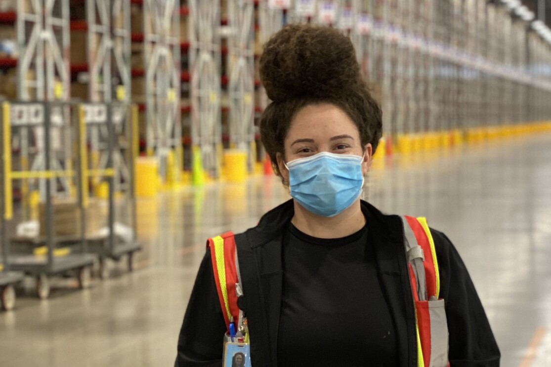 A woman wearing a safety vest and a mask stands in a warehouse space.