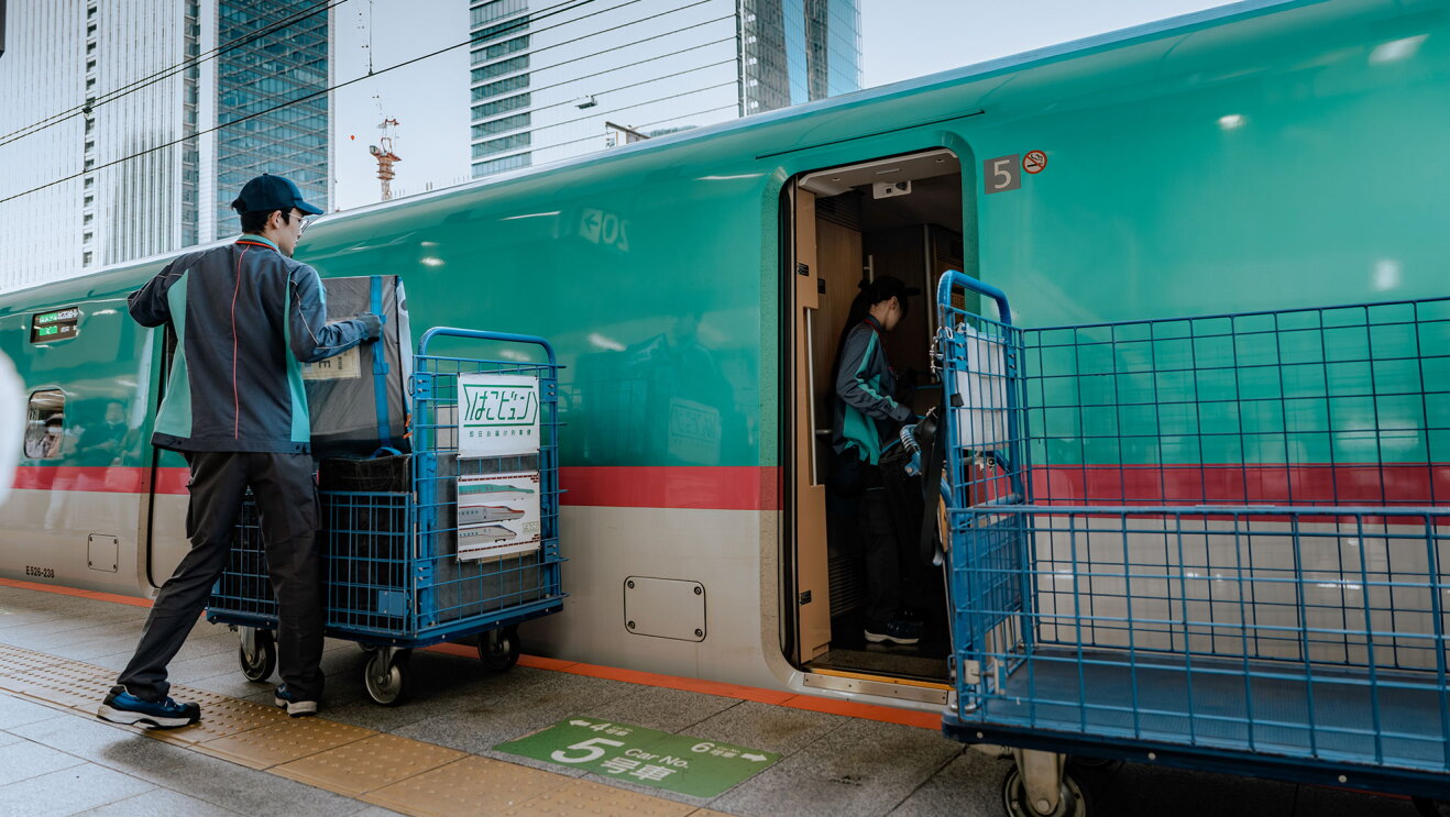 新幹線が停車中のプラットフォームで、配送員が台車から搬送用ボックスを下ろしている。新幹線のドアが開いていて、もう1人の配送員が車内にいる。手前にからの台車。