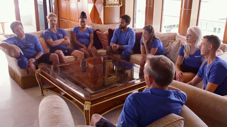 The crew in Below Deck gather in matching blue uniforms around an ornate coffee table
