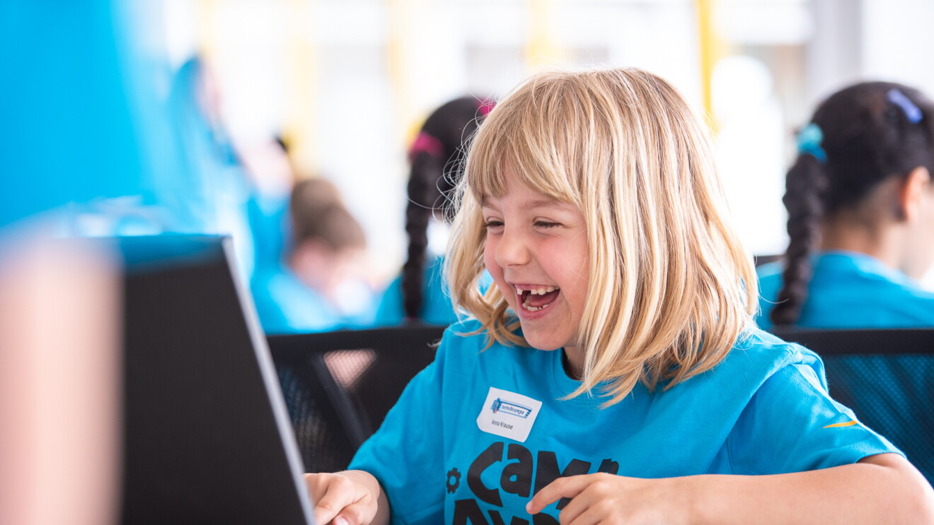 Ein blondes Mädchen im blauen Camp-Amazon-Shirt sitzt vor einem Laptop und lacht