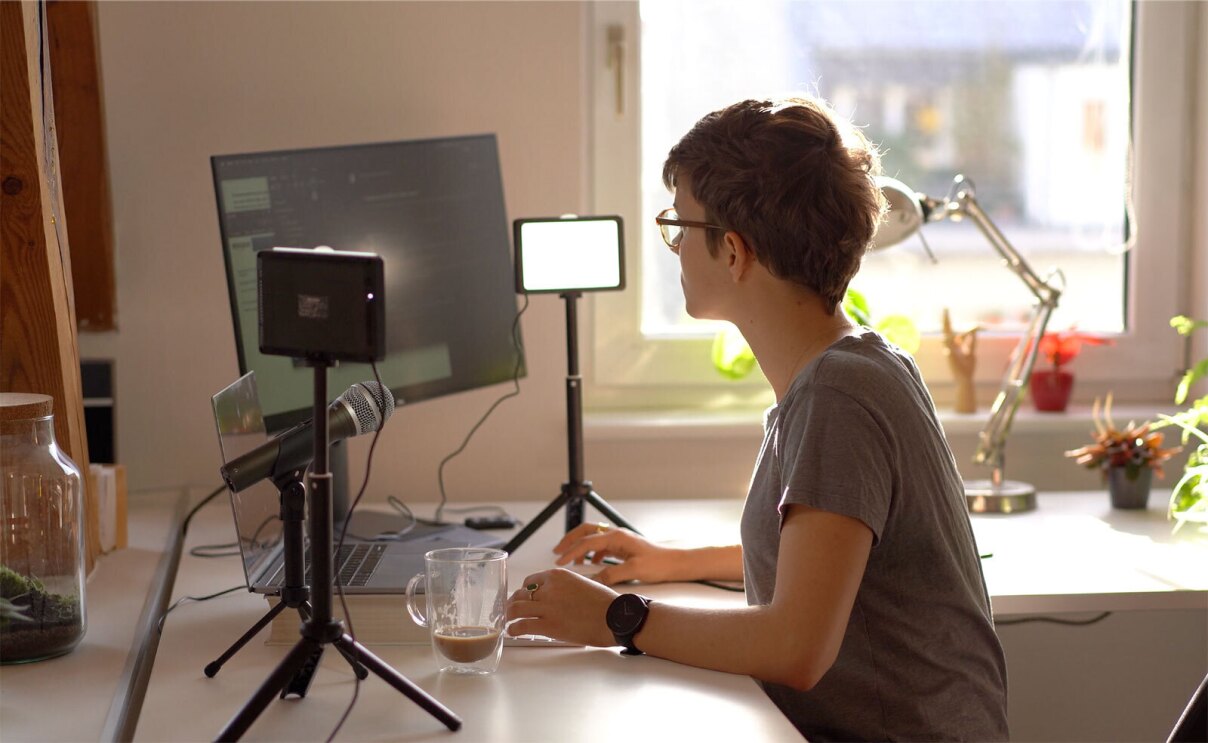 A professional photo of Antonia sitting at her desk with recording equipment surrounding her and an iced coffee in front of her.