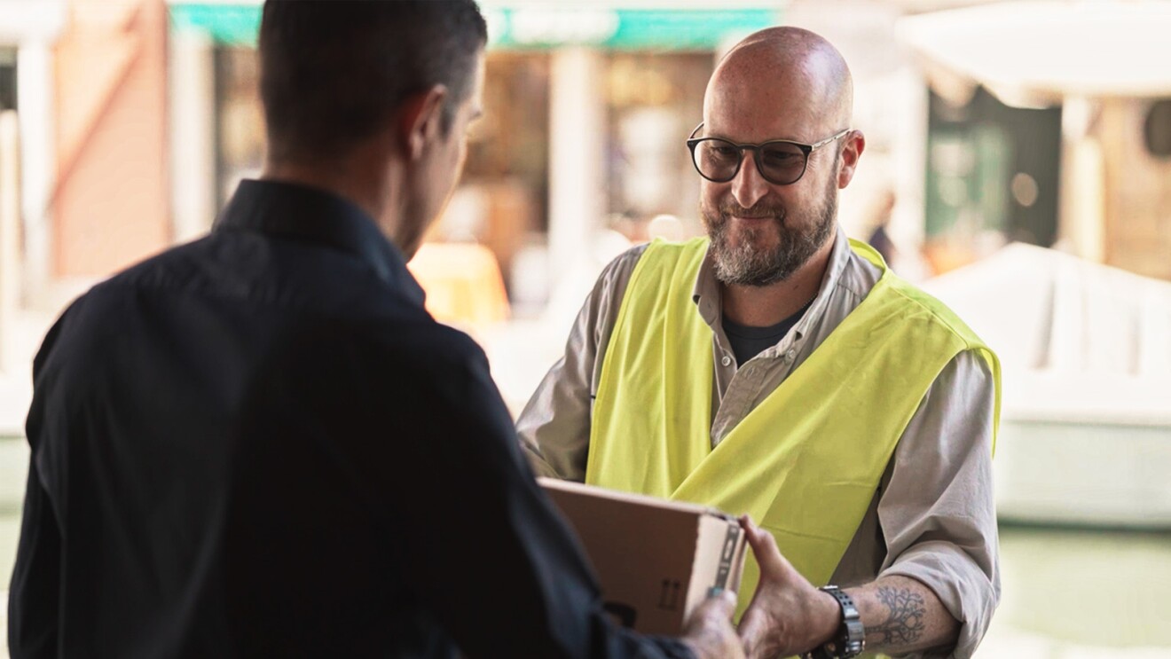 Roberto wears a yellow safety vest and delivers a package to Simone at his doorstep.