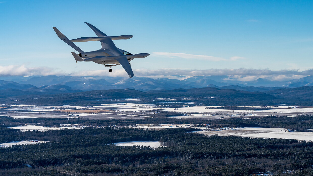 De fondo hay unas montañas nevadas y en primer plano bosque con árboles frondosos y muchos lagos. En primer plano un avión de BETA Technologies de color blanco con una parte inferior como si fuera un helicóptero y por encima unas alas con forma de avión de papel. Se le ve dos ruedas de aterrizaje. 