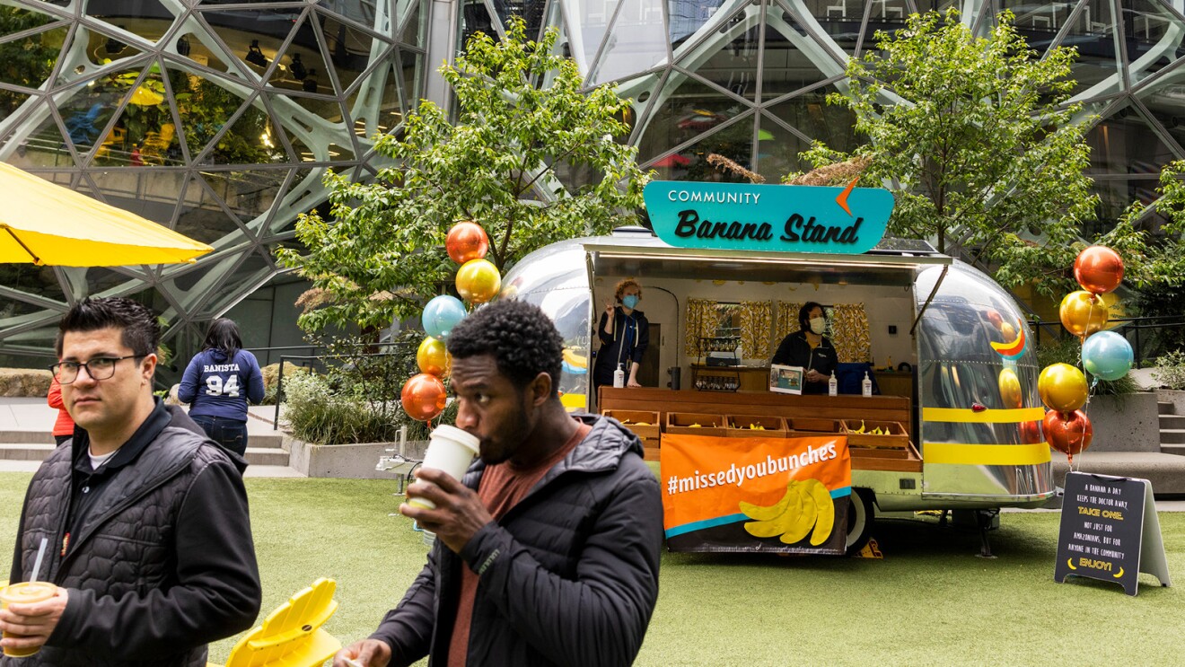 One of Amazon’s community banana stands sits outside the Spheres, Amazon’s urban rainforest in downtown Seattle. The left side shows two employees sipping coffee. 