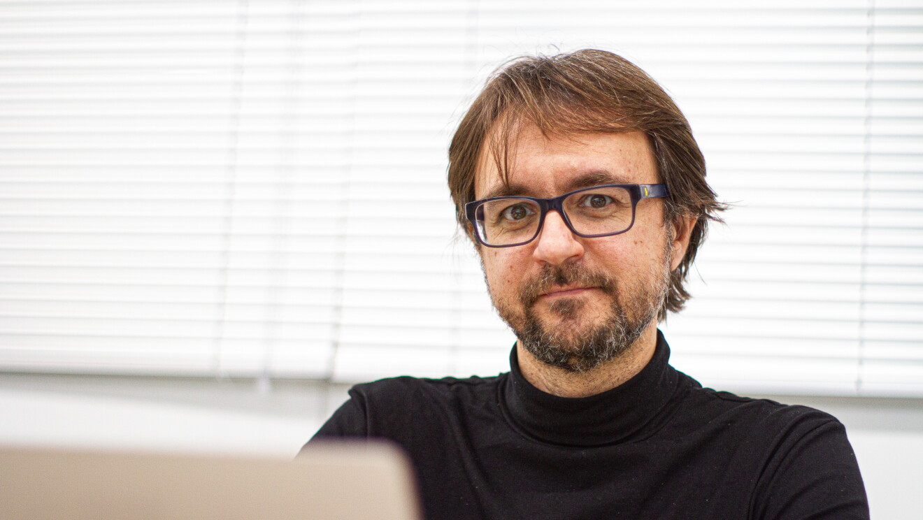 Hugo Zaragoza, investigador del Amazon Search Science y AI Group. Está en la oficina con unas cortinas detrás y un ordenador. Todo es de color blanco. ´Él lleva un jersey de cuello alto y unas gafas negras. Tiene barba y perilla de una semana y el pelo por las orejas. Estña mirando a cámara. Tiene barba negra con pelos blancos, y el pelo castaño.