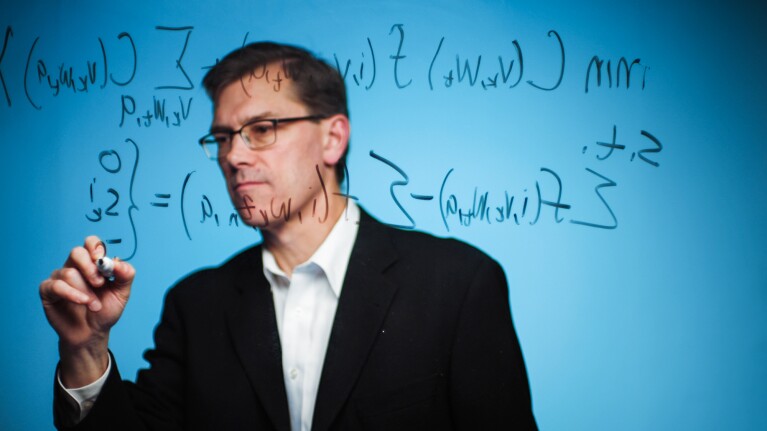 A man wearing a blazer and a button-down shirt writes a complicated equation.