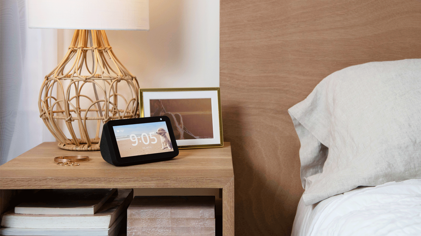 An image of an Amazon Echo Show device sitting on a wood night stand by a bed. There is a photo frame, a lamp, and jewelry next to the device.