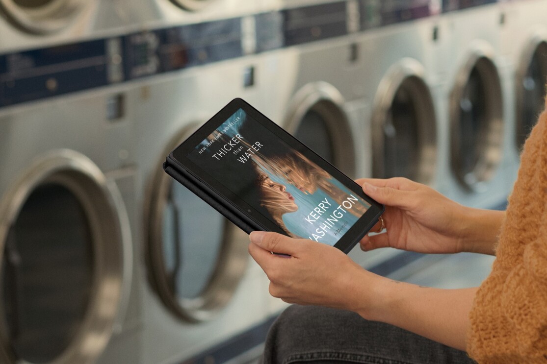 コインランドリーで黄色いセーターを着ている人は本の表紙が表示されているタブレットを持っている。