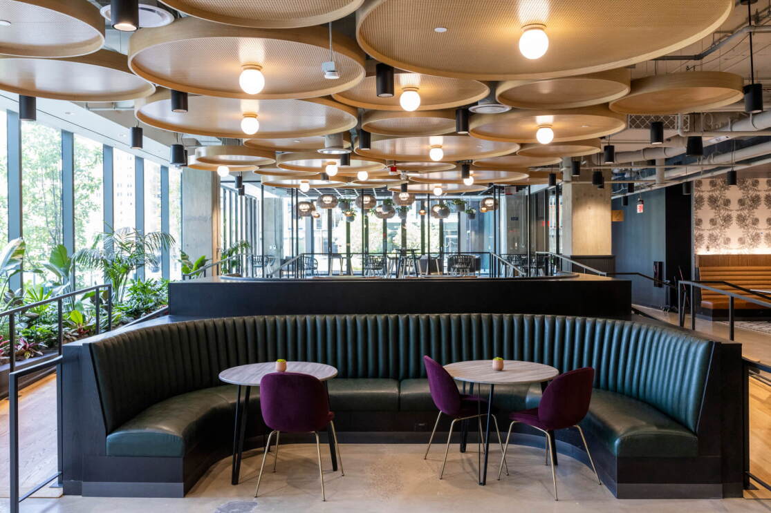 An image of a large dining booth with teal leather. There are large, tan light fixtures on the ceiling about and another booth behind it, and large, tall windows on the walls. 