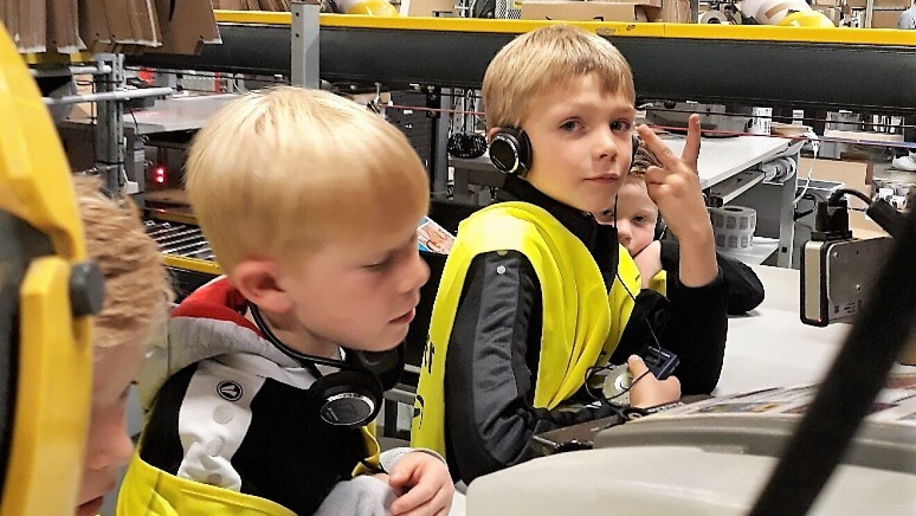 Zwei kleine Jungs in gelben Sicherheitswesten an einem Packtisch: Einer macht das Siegeszeichen