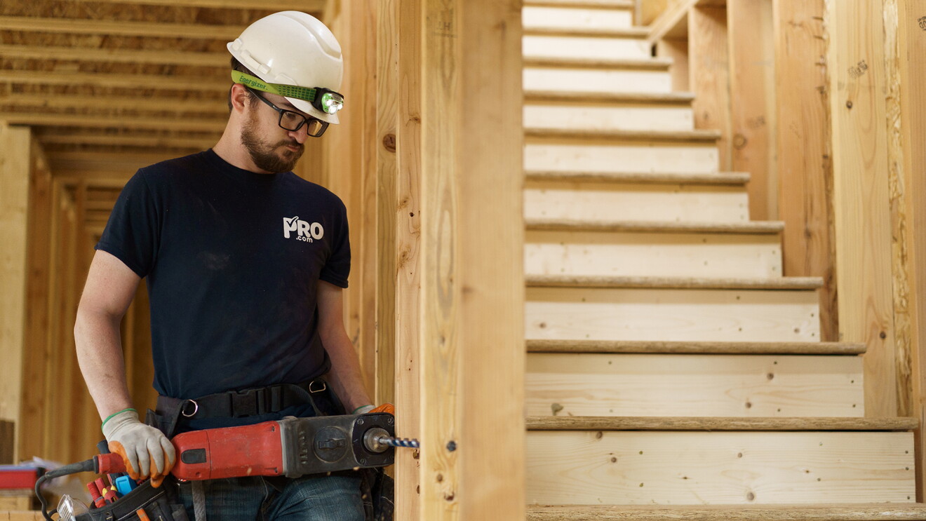 A male construction worker uses a drill next to a staircase. He wears a white hardhat, jeans, a tool belt, and a blue T-shirt with the Pro.com logo.