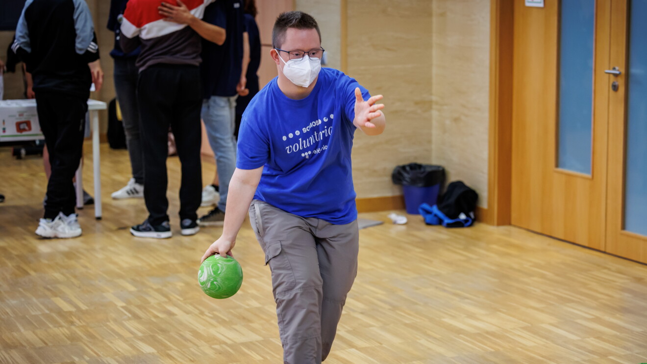 En las instalaciones de Down Madrid en Madrid aparecen 1 participante de la fundación con un bola de bowling agarrada y lista para tirarla contra los bolos. El participante va vestido con una camiseta azul y un pantalón gris. 