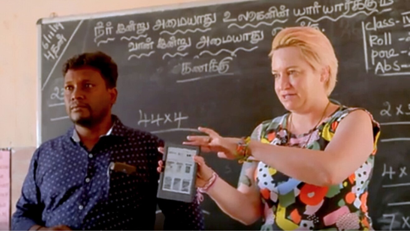 An image of two teachers in a classroom with a chalkboard behind them showing an Amazon Kindle device. 