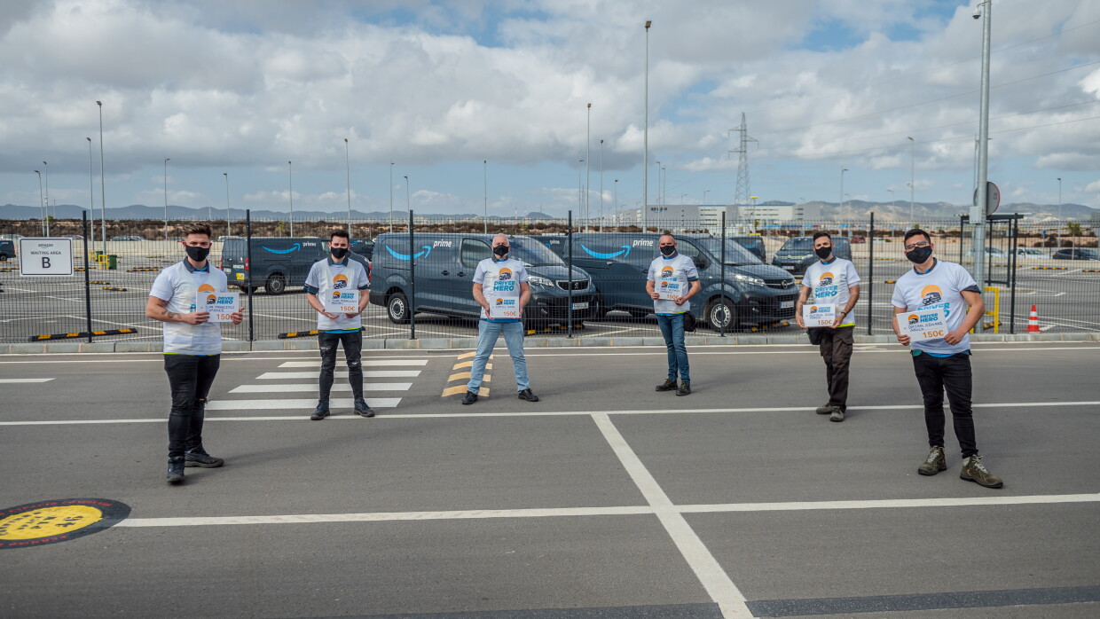 Foto de equipo con los reconocidos como Driver Hero. En un aparcamiento con las furgonetas de Amazon Prime de fondo están los 6 conductores reconocidos como Driver Hero. Todos ellos con una camiseta blanca y el logotipo del reconocimiento y en la mano un diploma. 
