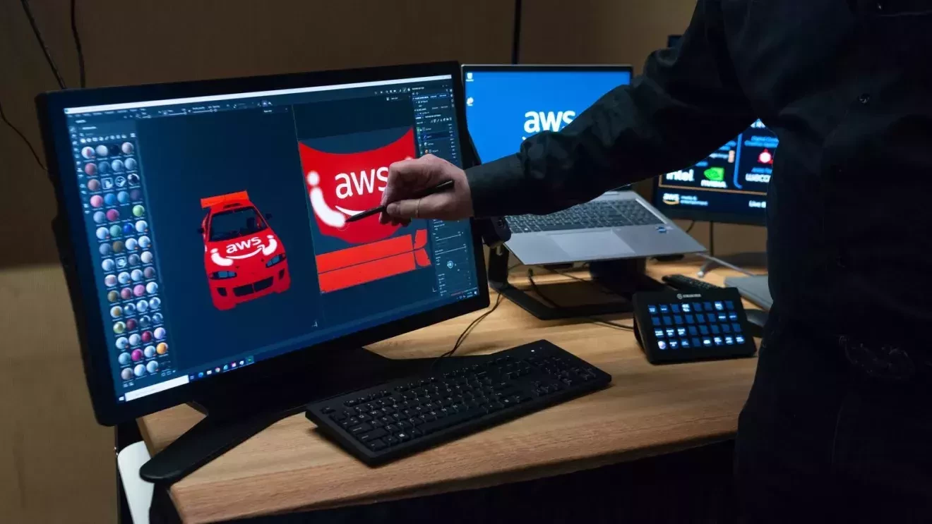 パソコン1台とモニター2台が机の上に置かれている。左にあるモニターには赤い車にAWSのロゴが書かれている。1人の手がそのモニターの中の赤い車に白い線を描いている。