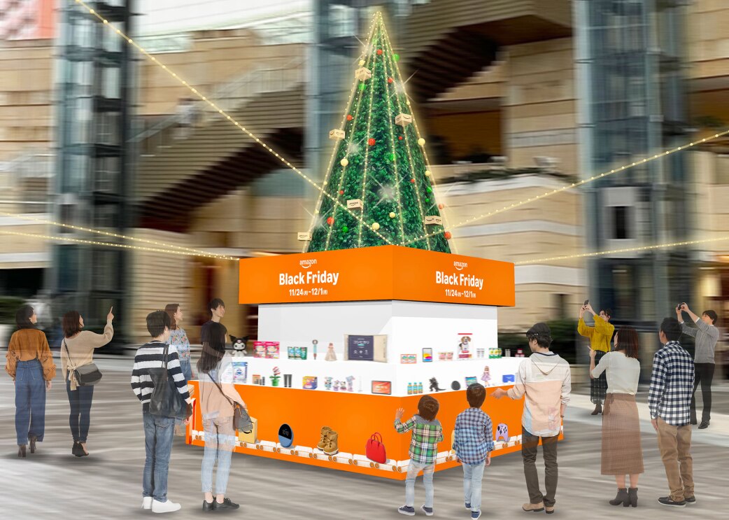 大規模施設の吹き抜けに設置されたクリスマスツリー。イルミネーションで装飾され、根元に、オレンジと白のパネルで作られたBlack Fridayの商品展示ブース。ツリーを見上げて指さしたり写真を撮る人々。