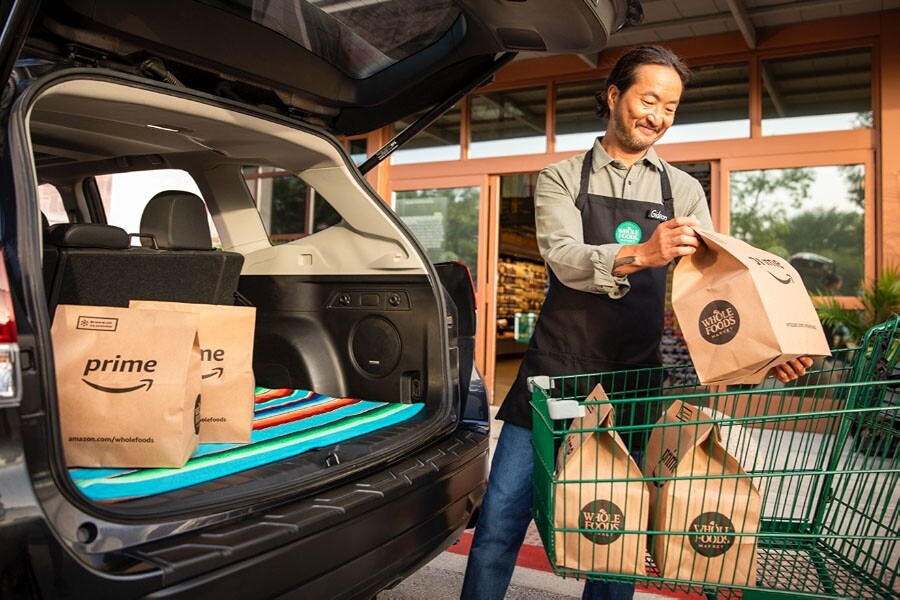 A Whole Foods Market employee loads grocery bags with the Prime logo on them in the back of a customer's vehicle.