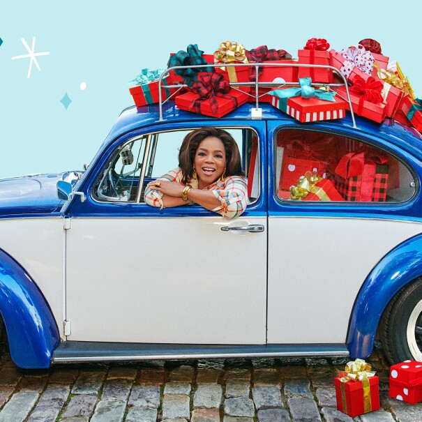 An image of Oprah smiling while looking out the window of a blue car. The car has presents stacked on top of and inside of it. 