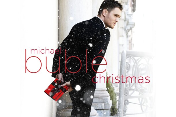 Michael Buble stands outside wearing a tuxedo, facing away, but seeming to be glancing over his shoulder. he holds a small, wrapped gift in red paper with black bow. Snow is falling in the image, and text saying "Michael Buble Christmas" is written over the top of the image.