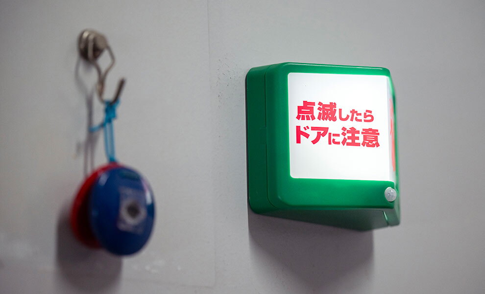 グレーの扉に設置された表示ランプ。緑色のフレームに囲まれた白い画面が光り、赤字で「点滅したらドアに注意」の表示。ランプの左側には青と赤のパーツから成るカスタネットがフックに掛けられている。