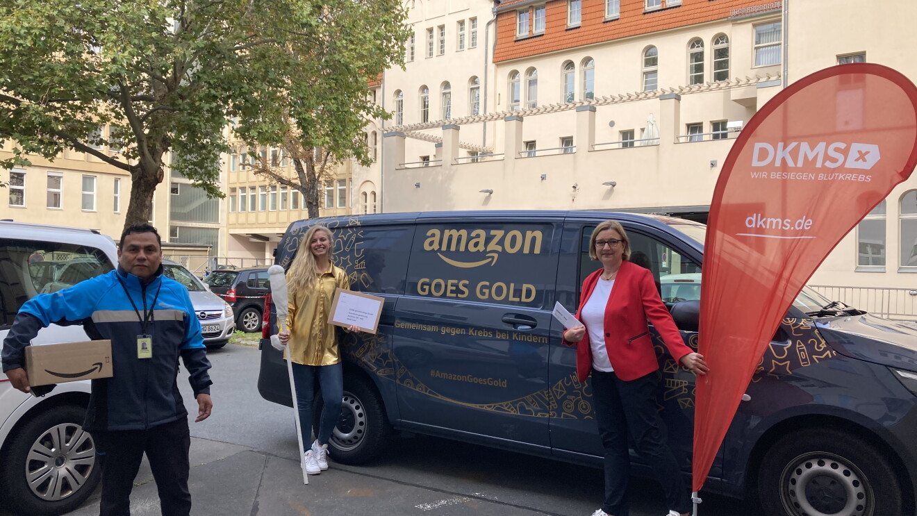 Amazon übergibt eine Spende an eine Vertreterin des DKMS. Die Vertreterin, eine Amazon Mitarbeiterin und ein Amazon Lieferpartner stehen vor einem mit "Amazon goes gold" beschrifteten blauen Liefervan.