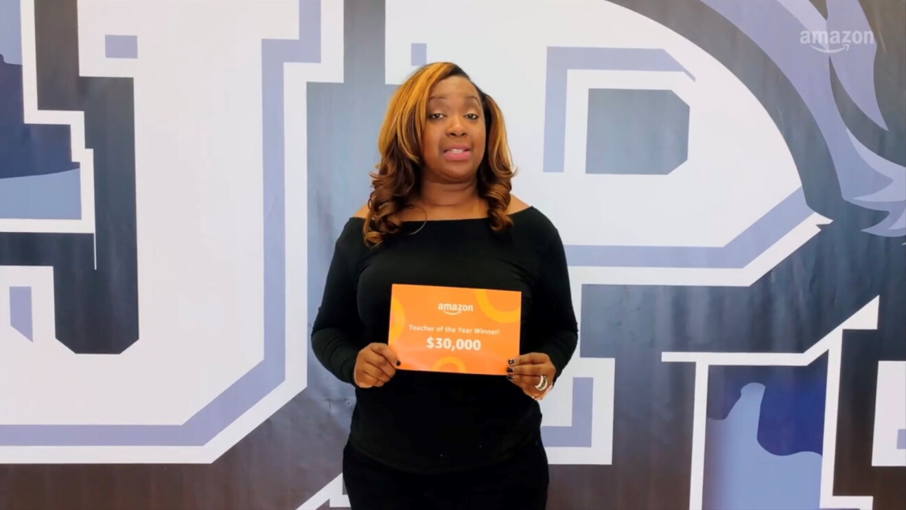 Melissa Collins wears a black dress and holds up an orange certificate that reads, "Amazon Teacher of the Year Winner $30,000." 