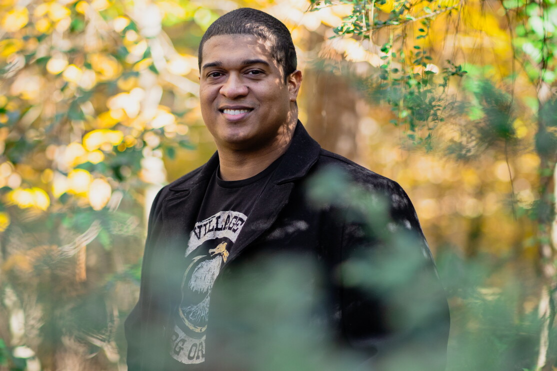 A man wearing a blazer and a T-shirt is photographed amidst foliage. Shown from the waist up, he has a short hair and smiles while looking directly into the camera.