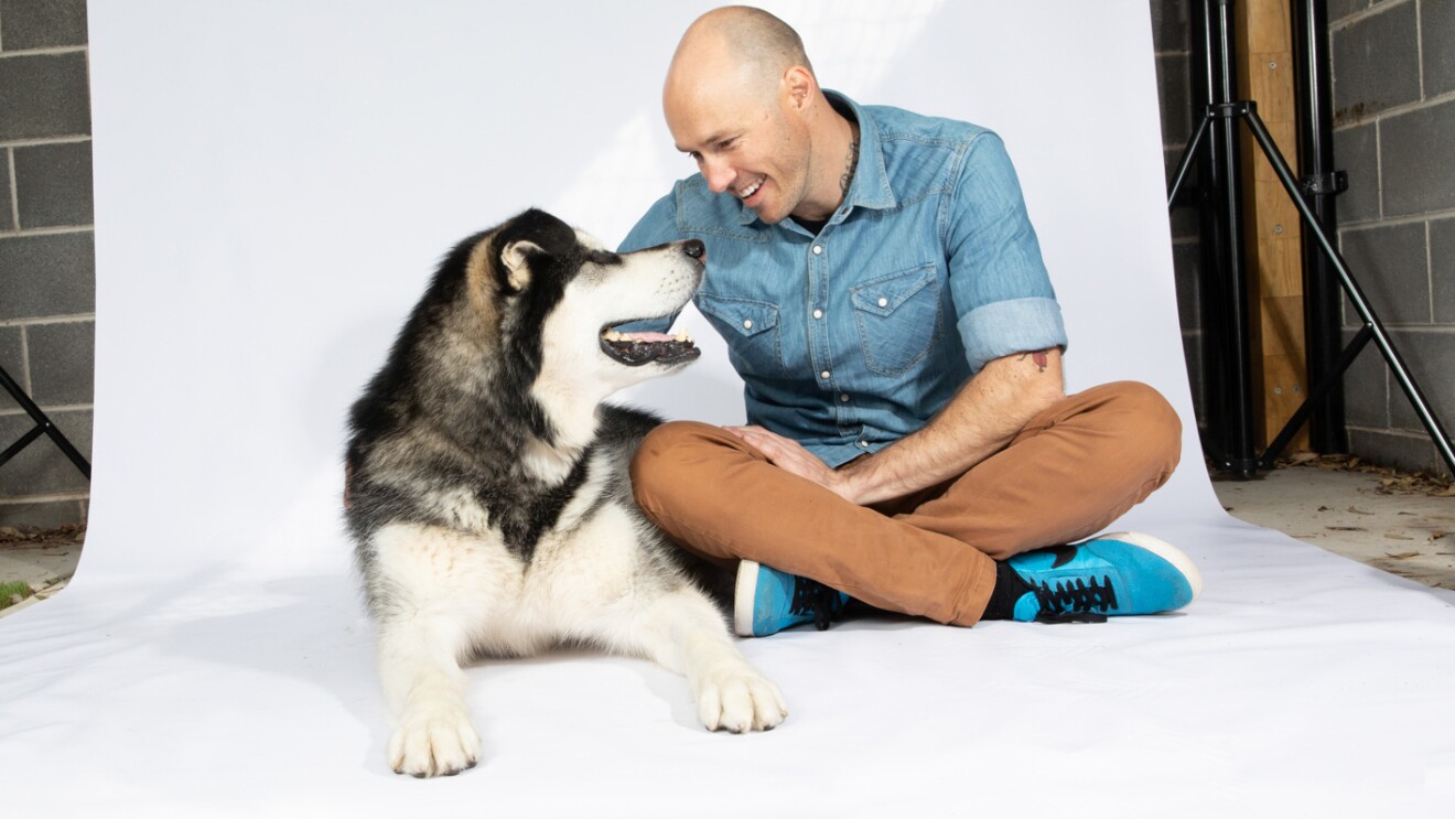 一名男子坐在白色背景上,穿着牛仔衬衫、卡其裤和网球鞋,俯下身去抚摸一只哈士奇狗,哈士奇狗抬头看着他。