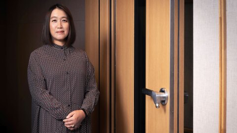 木製の分厚いドアの前に立つ人物。細かい幾何学模様のワンピースを着用し、両手を体の前で組んでいる。明るい室内で、人物の背後だけやや薄暗い。