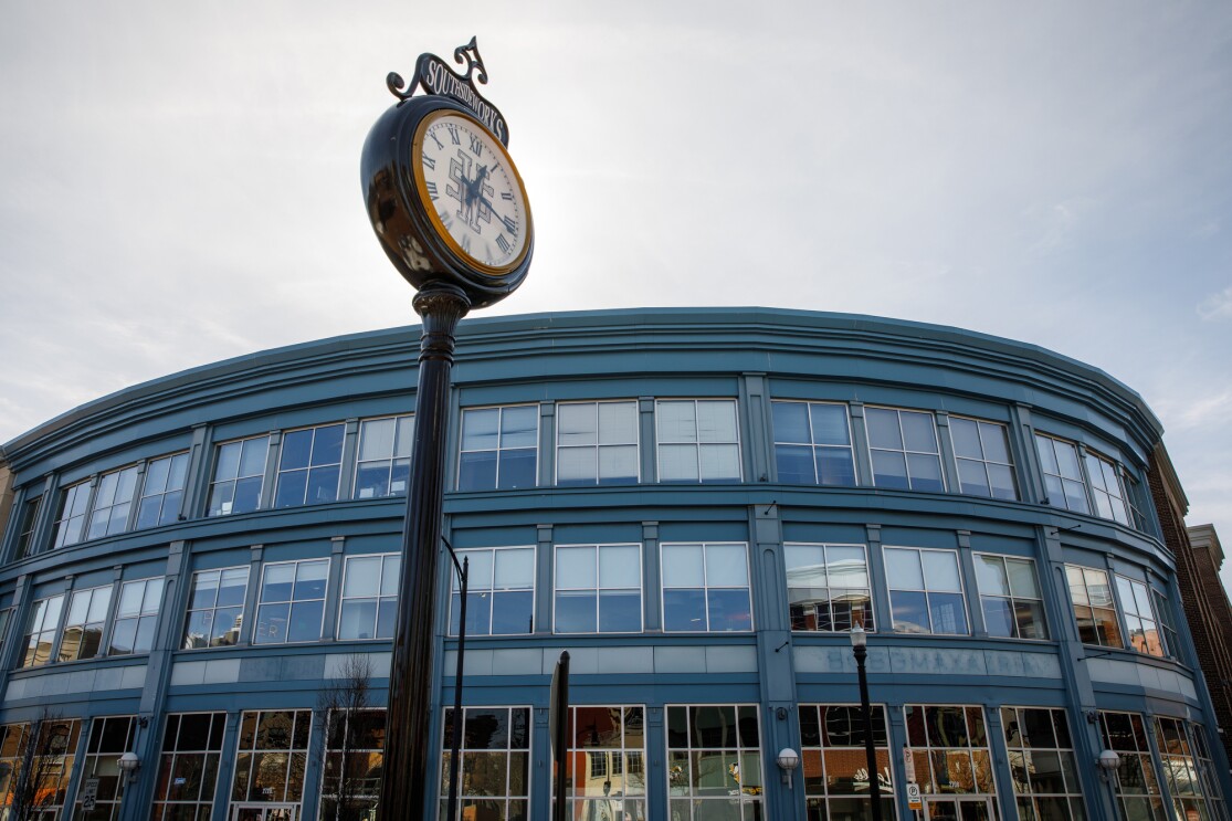 A view of the Amazon Pittsburgh office from the street. The building shows a wall of windows, three stories high. In front of the building is a clock on a light post.