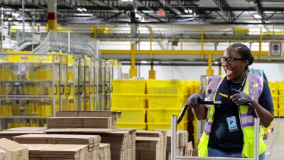 A Black female Amazon employee pushes a cart with boxes on it while smiling at somebody in an Amazon fulfillment center