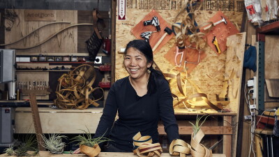 A woman stands behind a work table, smiling at the camera. In front of her are several handmade bentwood air plant containers, behind her sits more wood and tools. 
