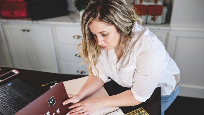 一个女人穿着白色纽扣和牛仔裤,趴在一本书的面料色板。她的右边是一台电脑和手机。在她身后是内置橱柜。