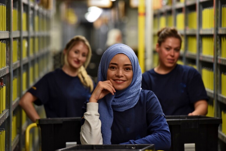 3 Logistikmitarbeiterinnen stehen in einem Regalflur eines Logistikzentrums. Alle drei schieben Kartwagen für die im Logistikzentrum gelagerten Produkte. Die Mitarbeiterin in der Mitte trägt ein Kopftuch, sie lächelt, stützt einen Arm auf den Wagen. Ihre beiden Kolleginnen sind unscharf im Hintergrund zu sehen, sie lächeln ebenfalls.