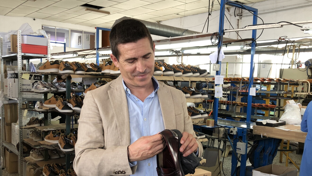 José Antonio Mollá, fundador de Castellanisimos, en una de sus fábricas, observando un zapato castellano que tiene en sus mannos. Detrás hay cahas de zapatos y una persona trabajando en una mesa. 