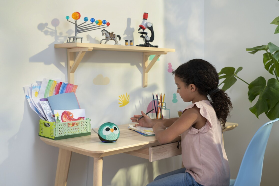 Ein Mädchen sitzt am Schreibtisch und malt in ein Malbuch. Neben ihr steht der neue Amazon Echo Dot Kids.
