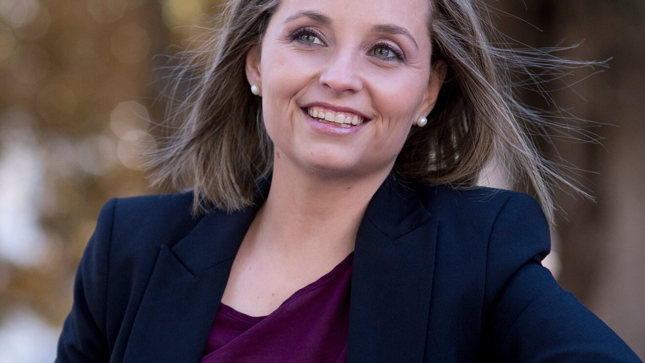 Almudena Navarro sonriendo lleva una americana azul y un jersey morado. De ojos azules y con media melena, el viento le mueve el pelo y deja ver sus pendientes.
