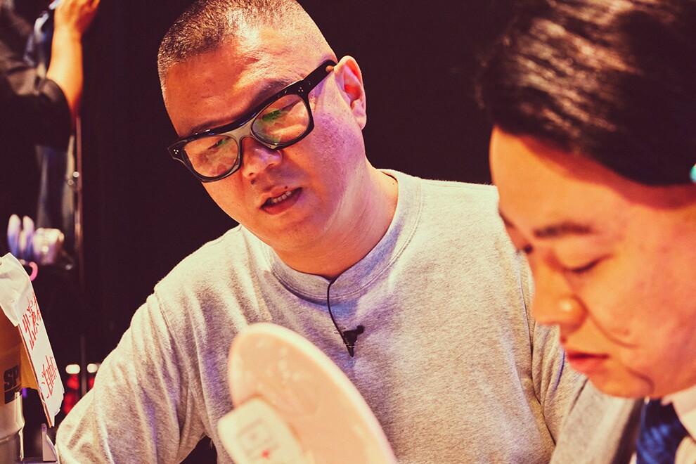 黒い壁を背に立つ2人の男性のクローズアップ。左側の男性はグレーのTシャツ、黒ぶち眼鏡。右側の男性は顔だけが写っている。2人とも真剣な表情。