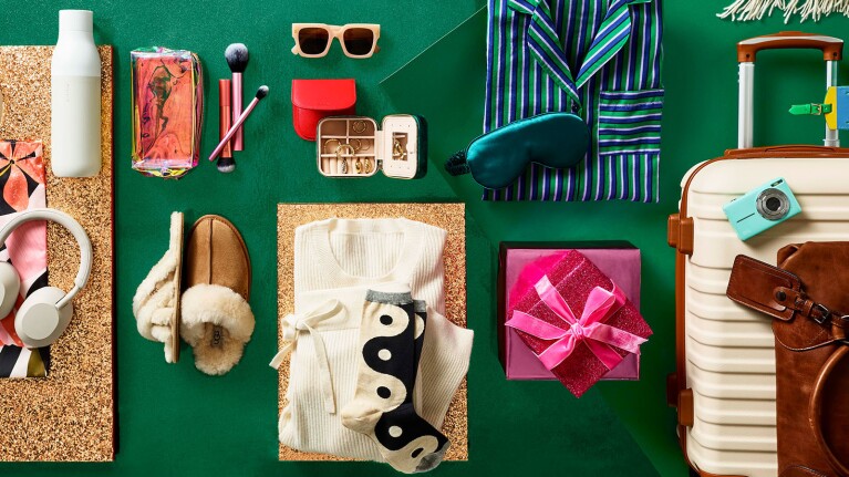An image of various clothing items and accessories against a green background. There is a sweater, socks with a yin-yang design, a blue sleep mask, white headphones, fuzzy slippers, makeup brushes, a water bottle, striped pajamas, an small jewelry box with rings in it, a blue camera, and a luggage set. There is also a pink wrapped gift.