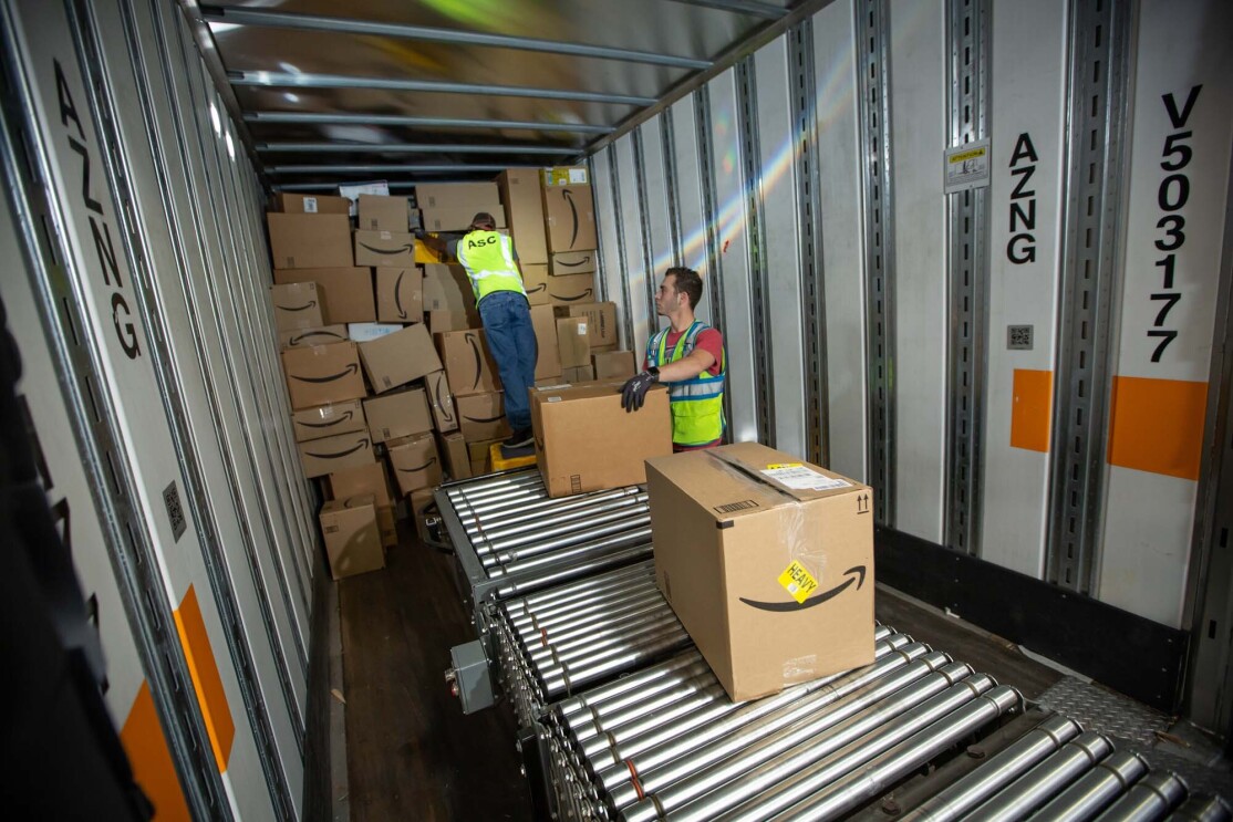 Two Amazon associates receive packages from a conveyor belt, as they prepare to place them on a trailer for distribution at a Columbus, Ohio fulfillment center, CMH2