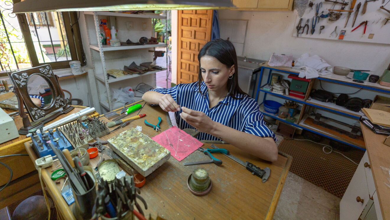 Sonia Llamas tiene 28 años y hace tres creó Iyé Biyé, su propio negocio online de joyería. Con el pelo largo castaño, lleva una camisa de rayas blancas y azules y se encuentra sentada en una mesa de su taller y manipulando un collar. La mesa está llena de herramientas de joyería y en el fondo también se ven muebles con herramientas incluso colgadas en la pared. En primerísimo primer plano, en la parte superior hay un foco para trabajar. 